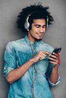Loving that song. Cheerful young African man wearing headphones and using smartphone with smile while standing against grey background photo