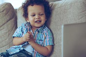 Funny cartoon. Cheerful little African baby boy looking at his laptop with smile while sitting on the couch at home photo