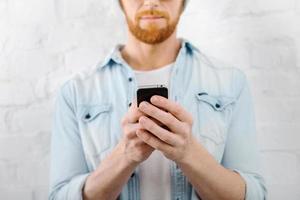 hombre enviando mensajes de texto. imagen recortada de un hombre barbudo sosteniendo un teléfono móvil mientras está de pie contra una pared de ladrillos foto