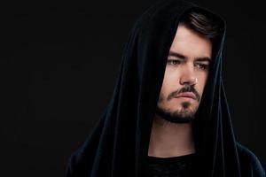 Handsome man in black. Pensive young man in hood looking away while standing against black background photo