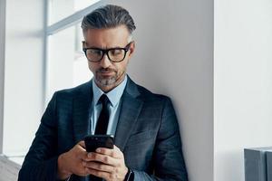 Confident mature man in formalwear using smart phone while standing near the window in office photo