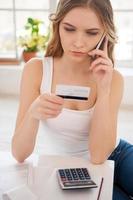 Financial problems. Top view of serious young woman holding credit card and talking on the mobile phone while sitting on the floor at home photo