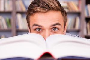 Just inspired. Excited young man looking out of the book while sitting against bookshelf photo