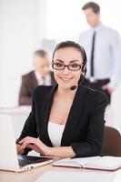 representante de servicio al cliente en el trabajo. mujer joven atractiva en ropa formal ajustando sus auriculares y sonriendo mientras la gente trabaja en segundo plano foto