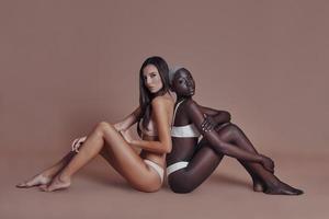 True feminine beauty. Two attractive mixed race women looking at camera while sitting back to back against brown background photo