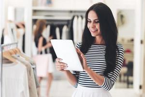 Technologies make business easier. Beautiful young woman using her digital tablet with smile while standing at the clothing store photo