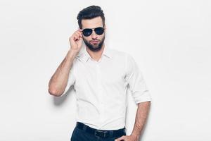 verdadero rompecorazones. un joven apuesto con camisa blanca ajustando sus gafas de sol y mirando a la cámara mientras se enfrenta a un fondo blanco foto