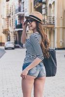 Casual beauty.  Rear view of beautiful young woman with bag over shoulder looking away and smiling while walking along the street photo