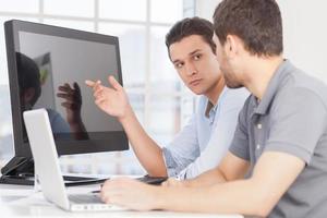 trabajando en el proyecto. dos jóvenes confiados discutiendo algo y señalando el monitor de la computadora mientras están sentados en la oficina foto