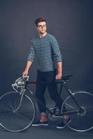 His bike is his lifestyle. Full length of confident young handsome man in glasses leaning at his retro styled bicycle and looking away while standing against grey background photo