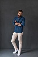 He knows everything about you. Full length of handsome young man looking at camera and keeping arms crossed while standing against grey background photo
