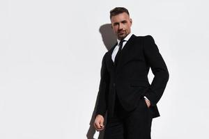 Used to look perfect. Handsome young man in full suit keeping hand in pocket and looking at camera with smile while standing against white background photo