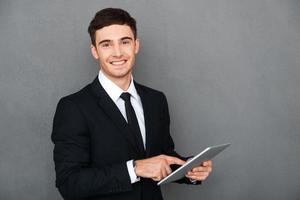 Businessman with digital tablet. Happy young man in formalwear working on digital tablet and looking at camera while standing against grey background photo