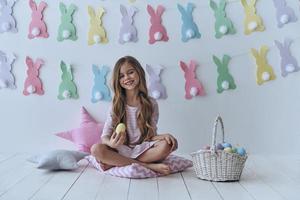 diversión de pascua linda niña sosteniendo un huevo de pascua y sonriendo mientras se sienta en la almohada con decoración en el fondo foto