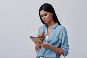 Thoughtful young woman holding note pad while standing against grey background photo