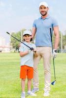 Favorite family game. Cheerful young man embracing his son while standing on the golf course photo