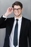 Confident businessman. Handsome young man in formalwear adjusting his eyeglasses and smiling while standing against grey background photo