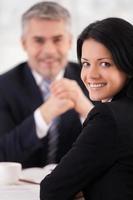 Woman on interview. Cheerful young woman in formalwear looking over shoulder and smiling while mature man in suit sitting on background photo