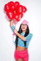 diversión con globos. feliz joven mujer africana con ropa funky sosteniendo globos de aire caliente en forma de corazón rojo y mirando hacia arriba con una sonrisa mientras está de pie contra el fondo blanco foto