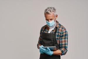 Senior man in apron and protective face mask using digital tablet while standing against gray background photo