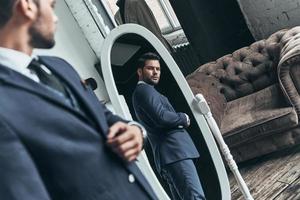 aspecto perfecto. reflejo de un apuesto joven con traje completo ajustando su chaqueta mientras está de pie frente al espejo en el interior foto