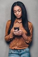 Message for him. Beautiful young African woman holding smart phone and looking at it while standing against grey background photo