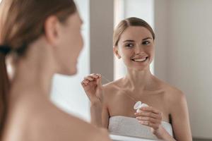 Good habit. Over the shoulder view of young beautiful woman holding dental floss and smiling while looking in the mirror photo