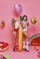 chicas supremamente sensuales. Longitud total de dos atractivas mujeres jóvenes en traje de baño tomando bebidas mientras están de pie contra el fondo rosa foto