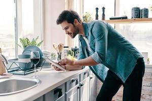 What to cook Handsome young man in casual wear using digital tablet and smiling while standing in the kitchen at home photo