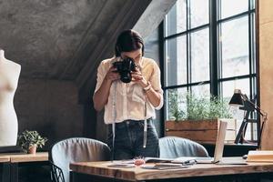 joven perfeccionista. mujer joven concentrada con cinta métrica en el cuello usando una cámara digital mientras está de pie cerca del escritorio en su taller