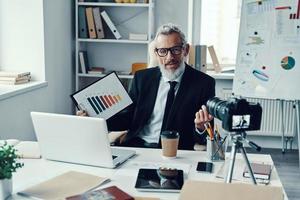 Confident mature man in elegant suit showing chart and sharing business experience while making social media video photo