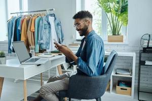 Confident young African man using smart phone while sitting in fashion store office photo