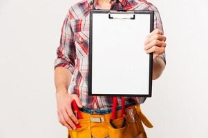 Copy space on his clipboard. Close-up of handyman with tool belt stretching out clipboard with paper standing against grey background photo