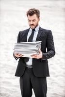 Tired of paperwork. Frustrated young businessman holding stack of paperwork while standing on sand in desert photo