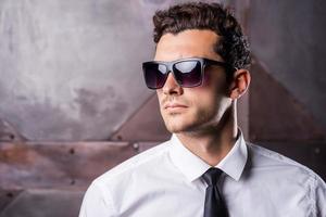 Stylish handsome. Handsome young man in formalwear and sunglasses looking away while standing against metal background photo