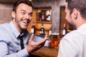 pasar la noche del viernes en el bar. dos jóvenes felices con camisa y corbata hablando entre ellos y gesticulando mientras beben cerveza en el mostrador del bar foto
