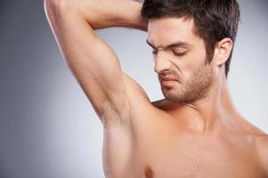 Unpleasant smell. Portrait of young shirtless man looking at his armpit and grimacing while standing isolated on grey background photo