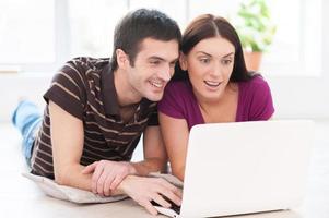 Spending their free time in the net. Beautiful young couple working on laptop together while lying on the floor at their apartment photo