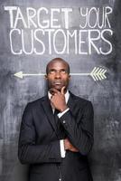 Target your customers. Thoughtful young African man in formalwear standing against blackboard with sketch on it and holding hand on chin photo