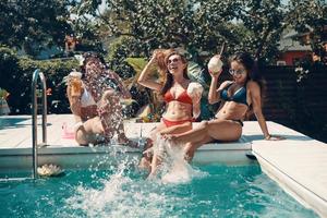 mujeres jóvenes atractivas en bikini bebiendo cócteles y sonriendo mientras se sientan junto a la piscina al aire libre foto