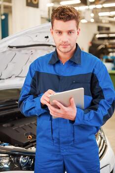 Concentrated on your problem. Confident young man working on digital tablet and looking at camera while standing in workshop with car in the background photo