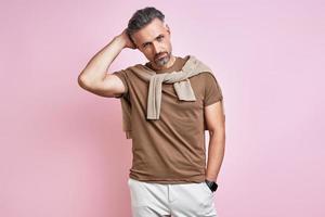 Handsome mature man holding hand behind head while standing against pink background photo