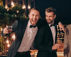 dos hombres guapos con traje bebiendo whisky y sonriendo mientras pasan tiempo en la fiesta foto