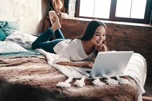 Enjoying lazy time at home. Beautiful young woman using laptop and smiling while lying on the bed at home photo