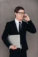 Staying in touch with the colleagues. Confident young man in formalwear carrying laptop and talking on the mobile phone while standing against grey background photo