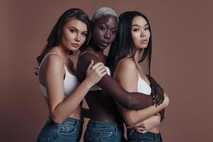 simplemente impresionante. tres mujeres jóvenes atractivas mirando a la cámara mientras están de pie contra el fondo marrón foto