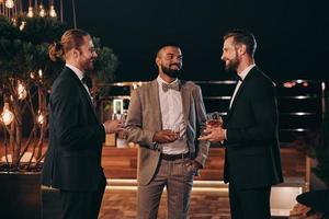 Three handsome men in suits drinking whiskey and communicating while spending time on party photo
