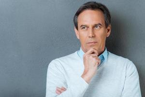 Lost in thoughts. Portrait of thoughtful mature man holding hand on chin and looking away while standing against grey background photo