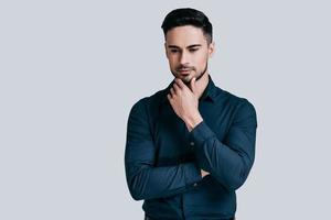 Thinking about solution. Good looking young man holding hand on chin and looking away while standing against grey background photo