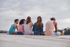 Young and carefree. Rear view of young people in casual wear smiling and talking while sitting on the pier photo